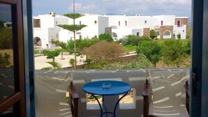 Ein Balkon mit zwei weißen Stühlen und einem kleinen runden Tisch, mit Blick auf ein weißes Gebäude mit Bögen und einen Garten.