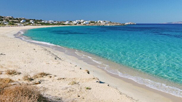 Ein Strand mit klarem türkisblauem Wasser, sandigem Ufer und weißen Gebäuden im Hintergrund.