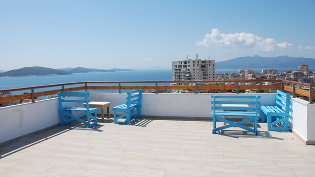 A rooftop terrace with blue chairs and a table, overlooking a city and the sea.
