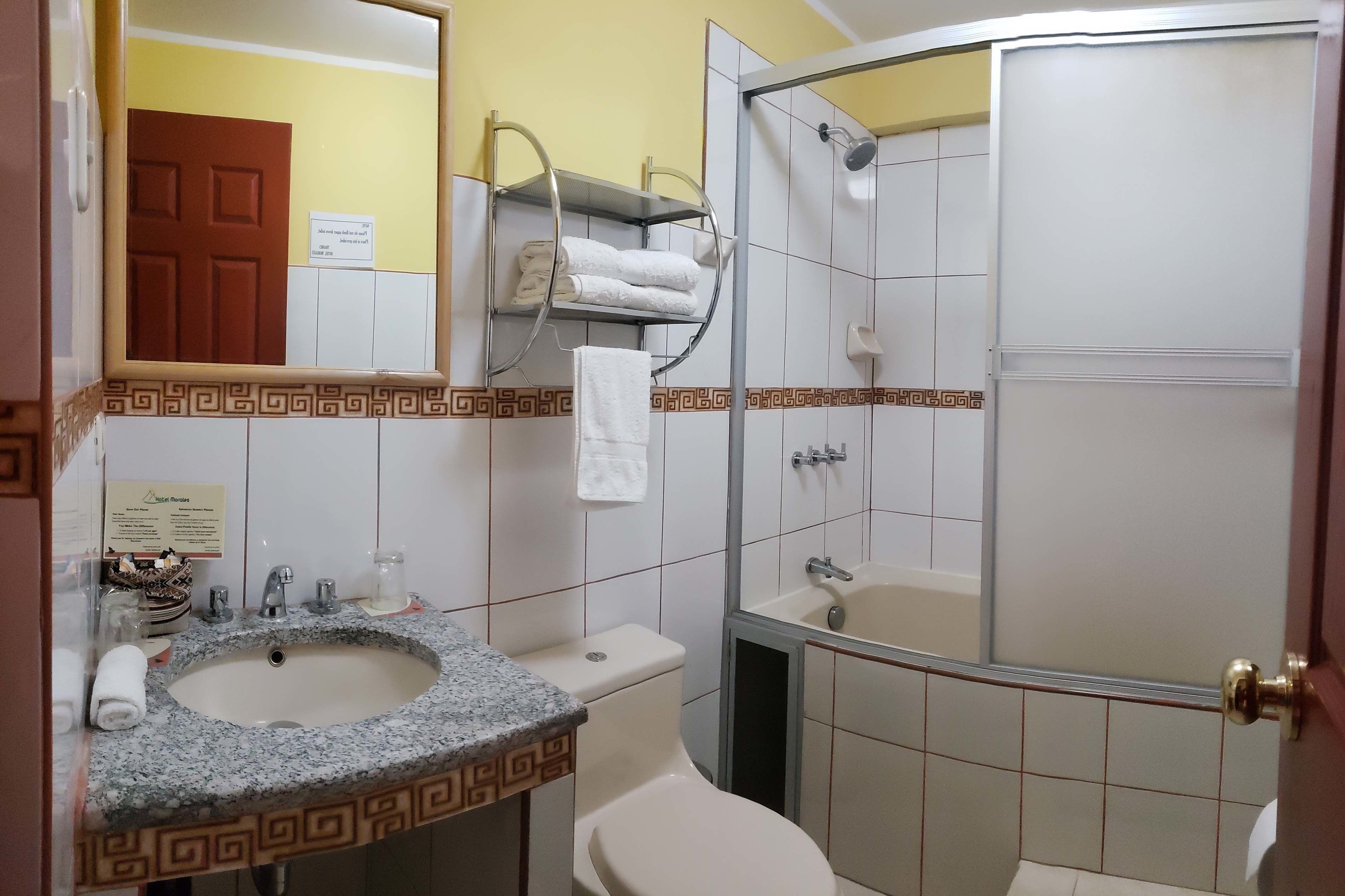 Un baño con lavamanos, inodoro y ducha. Hay un perchero con toallas, un espejo y una puerta.