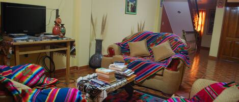Uma sala de estar com um sofá, poltrona e uma mesa de centro com livros. Há uma televisão em um móvel e um vaso na mesa de centro.