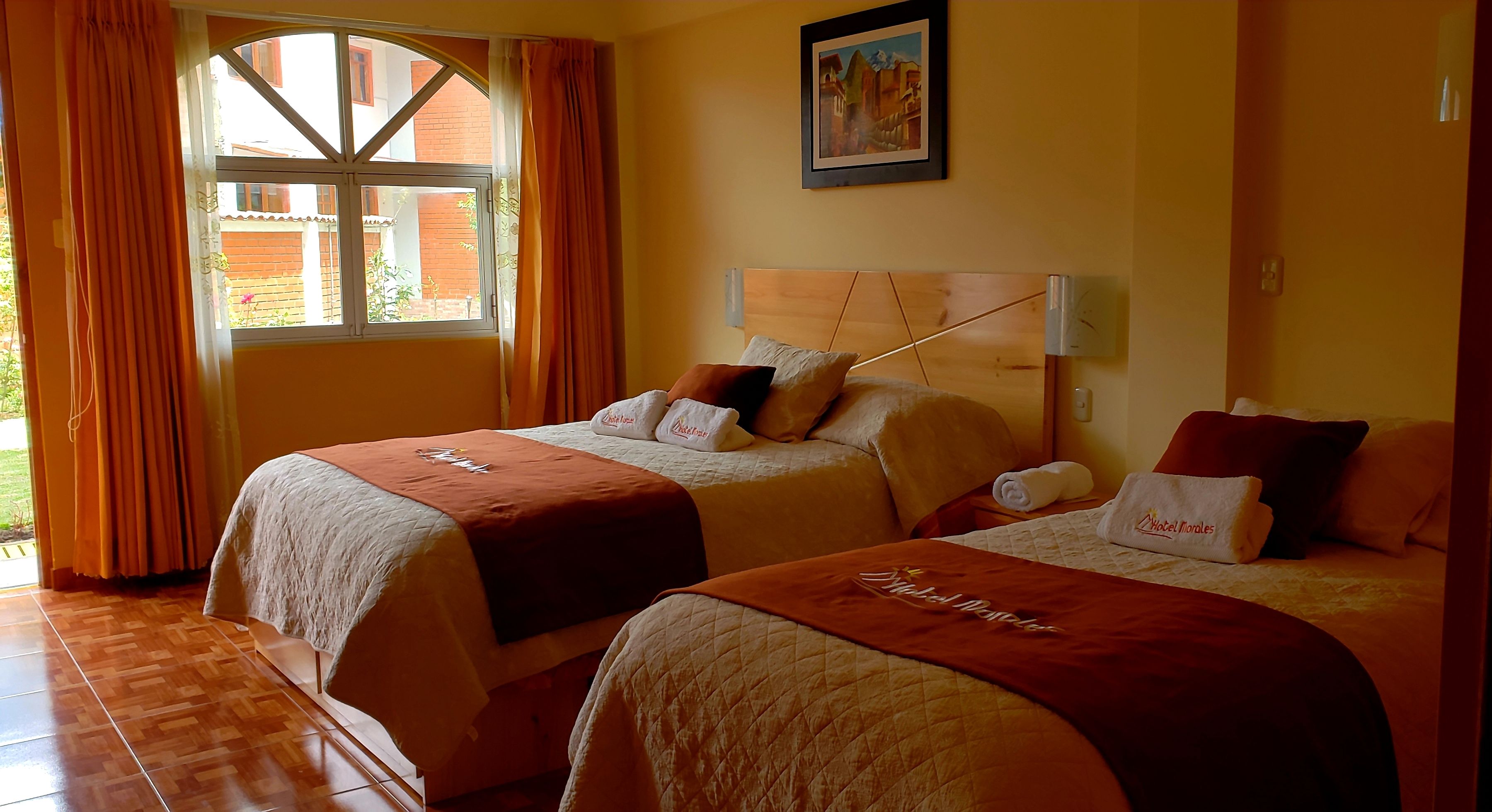 A hotel room with two beds, each with a white bedspread and a brown blanket. There is a window with curtains, a painting on the wall, and a wooden headboard.