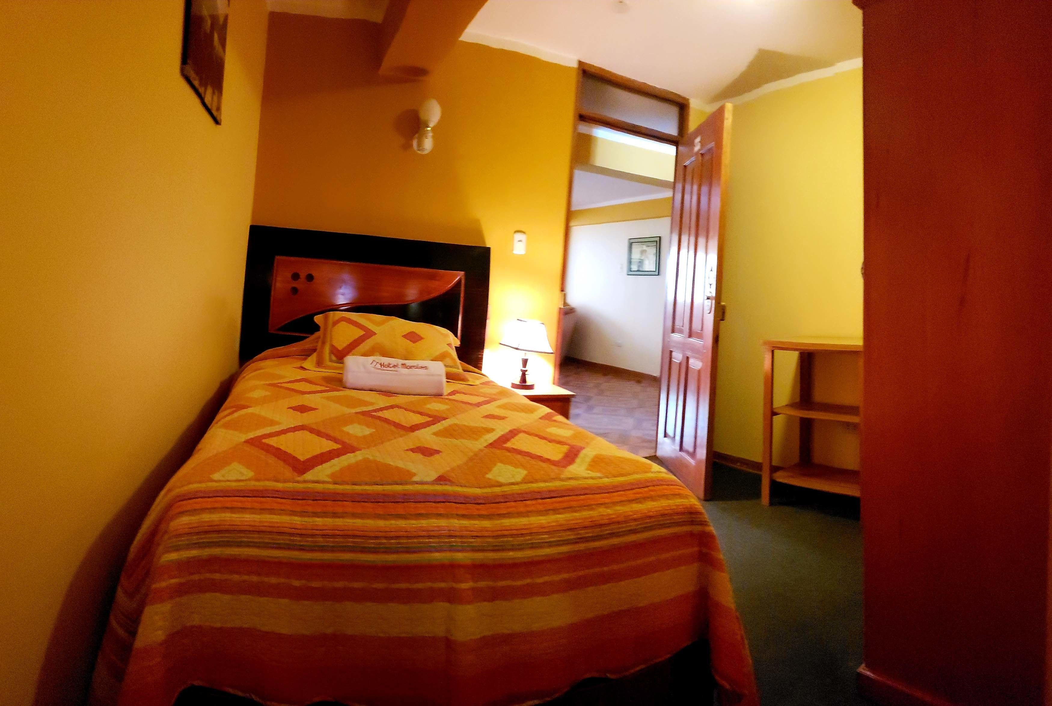 Een hotelkamer met een bed, nachtkastje, lamp en een deur naar een andere kamer.