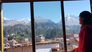 A person looking out of a window at a mountainous landscape with buildings in the foreground.