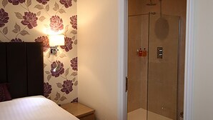 A hotel room with a bed, a nightstand, a wall-mounted lamp, and a glass-enclosed shower.