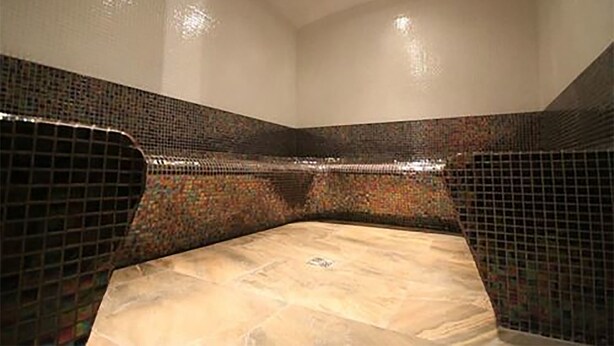 A room with a tiled floor and walls, a corner with a curved wall, and a ceiling with recessed lighting.