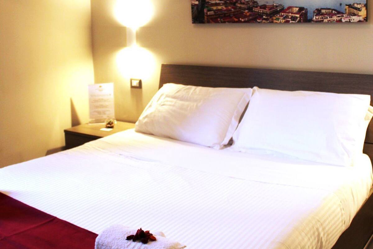 A neatly made bed with white linens, a red throw blanket, and a folded towel with a flower on it. There is a nightstand with a lamp and a framed picture on the wall above the bed.