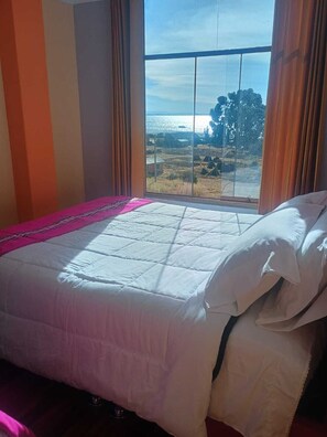 A bed with a white duvet and pink blanket, a window with curtains, and a view of a landscape with trees and a body of water. - Luquina homestay (Chucuito)