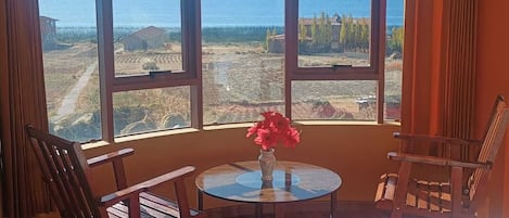 Una habitación con un ventanal grande que da a un cuerpo de agua y un paisaje, una mesa redonda con un florero y dos sillas de madera.