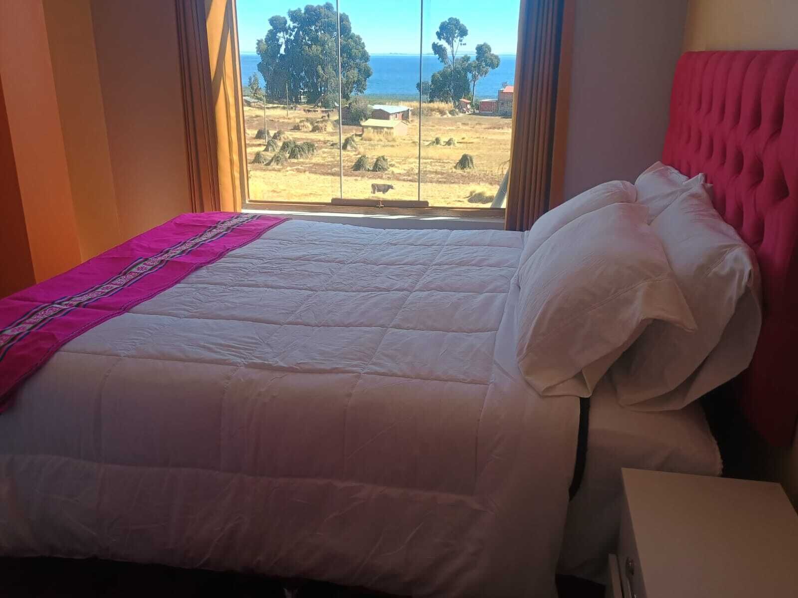 Cama con colcha blanca y detalles en rosa, ubicada cerca de una ventana con vista a un paisaje y árboles.
