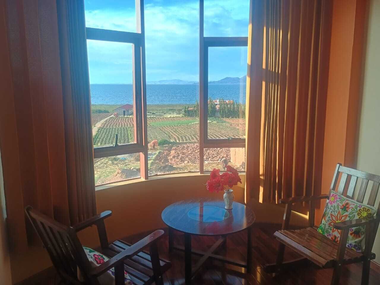 Una habitación con un ventanal amplio, dos sillas de madera, una mesita redonda con un florero y vistas a un paisaje de campos y montañas.