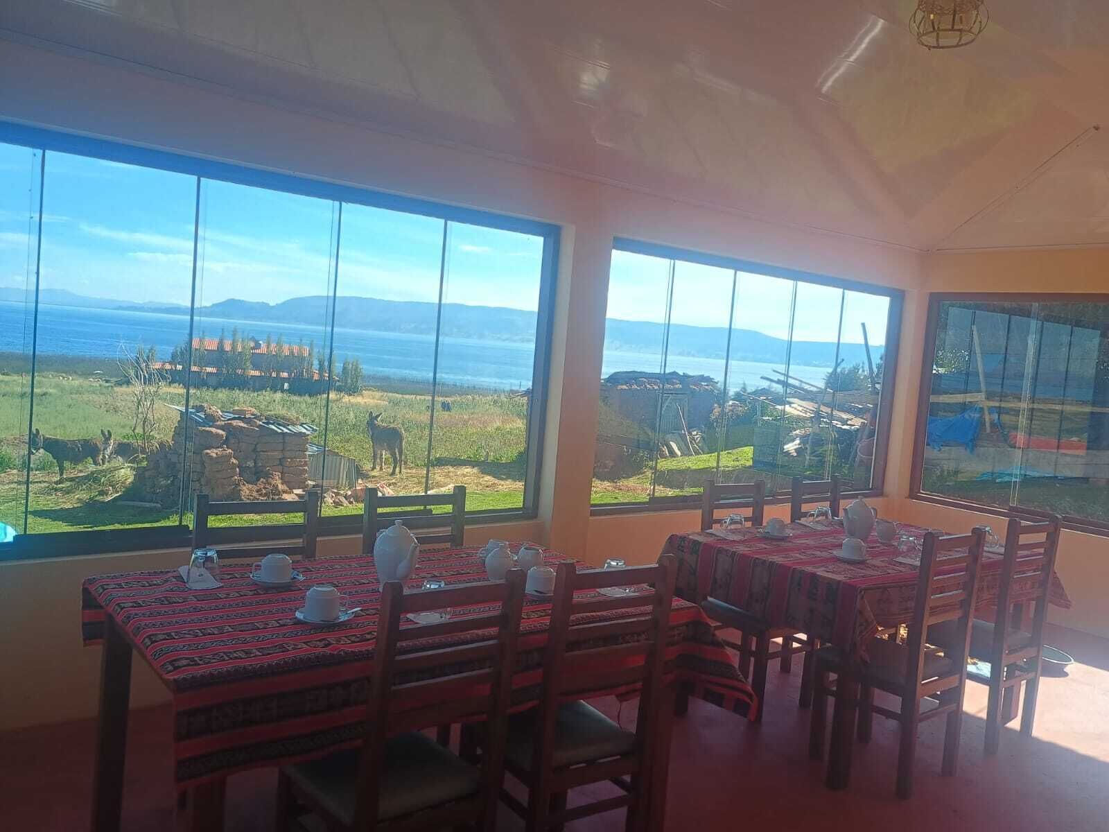 Un comedor con mesas y sillas, amplias ventanas que ofrecen vista a un paisaje con animales y una masa de agua.