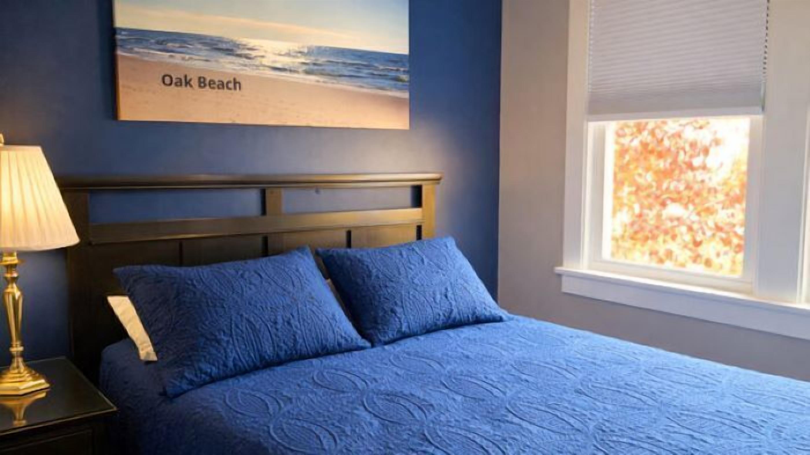 Un dormitorio con cama, mesita de noche, lámpara, ventana con persianas y un cuadro de una playa.