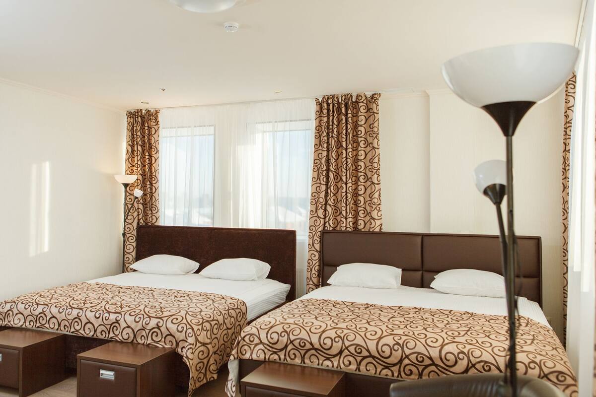 Hotellihuone, jossa on kaksi sänkyä. Jokaisessa sängyssä on kuviollinen päiväpeite ja samanväriset päätyosat. Huoneessa on yöpöydät laatikoilla, lattiavalaisin ja pyöriväkuvioidut verhot.