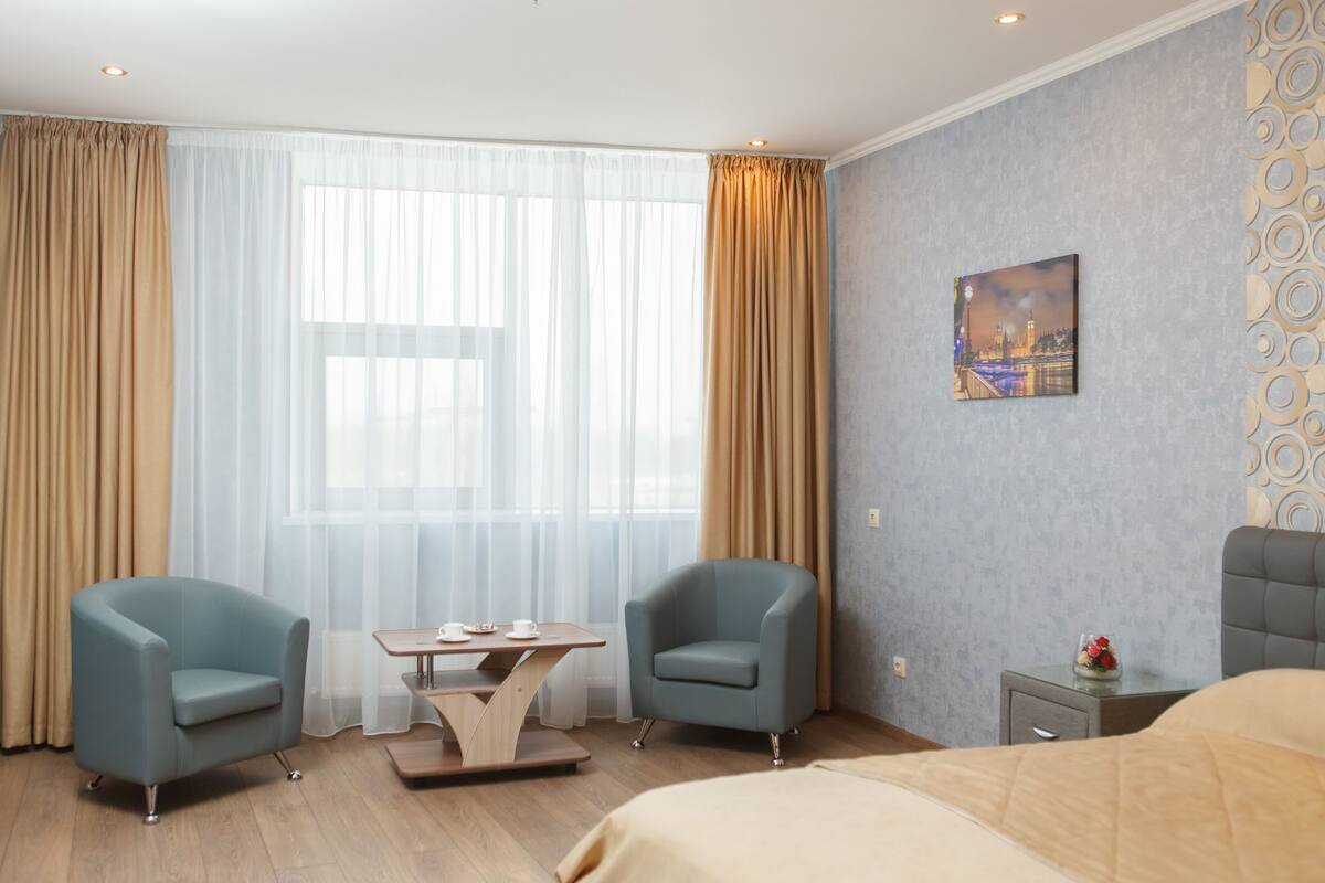 Hotellihuone, jossa on kaksi nojatuolia, puinen sohvapöytä, sänky, yöpöytä ja taulu seinällä.
