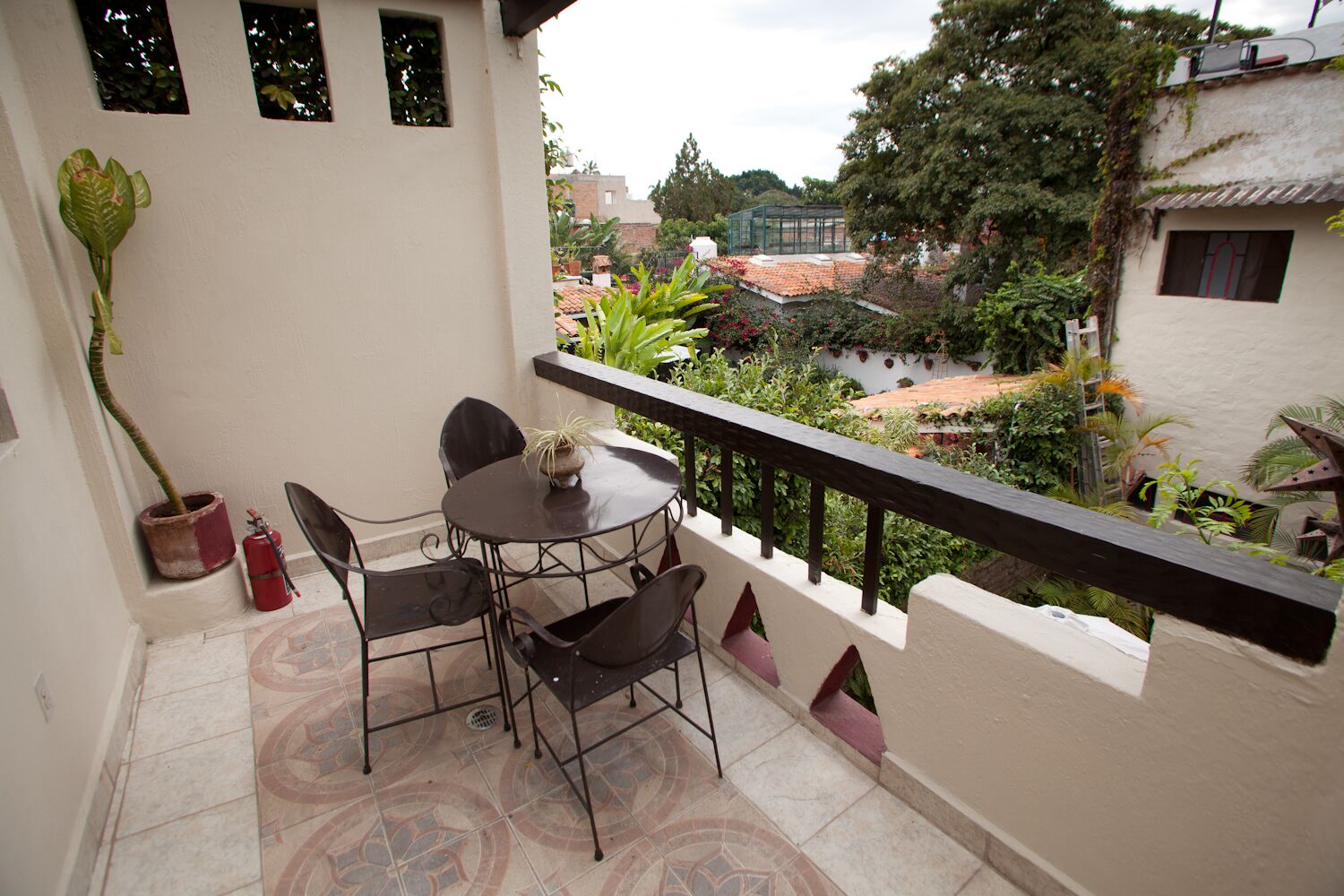 Uma varanda com mesa e cadeiras, um vaso de planta e vista para um jardim e edifícios.