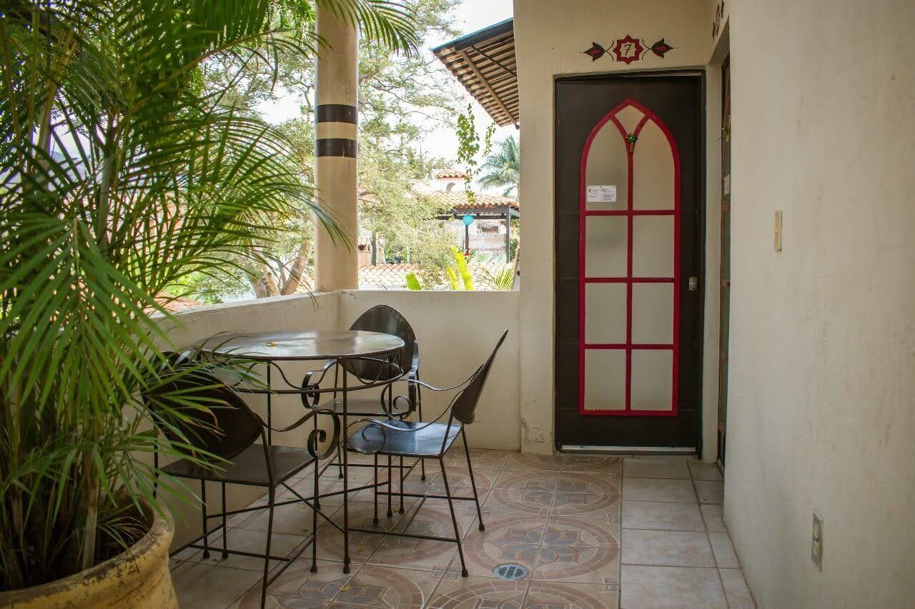 A small outdoor patio with a round table and chairs, a potted plant, and a door with a red frame.