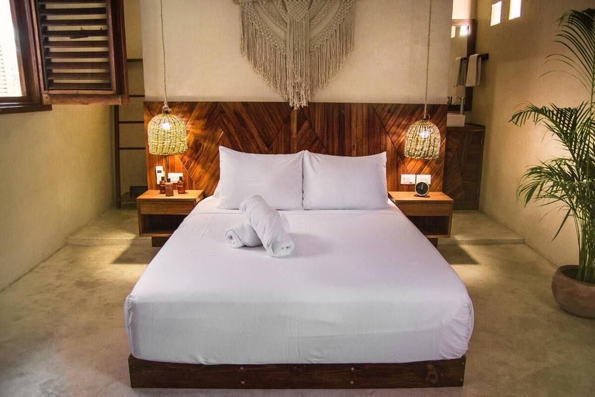 A neatly made bed with white linens and a rolled towel, wooden headboard, two bedside lamps, and a large plant to the right.