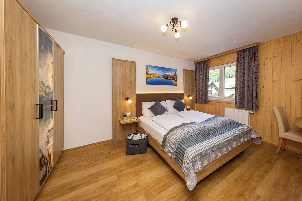 Quarto com piso de madeira, cama com manta estampada, mesa de cabeceira, luminária na parede, janela com cortinas e quadro sobre a cama.