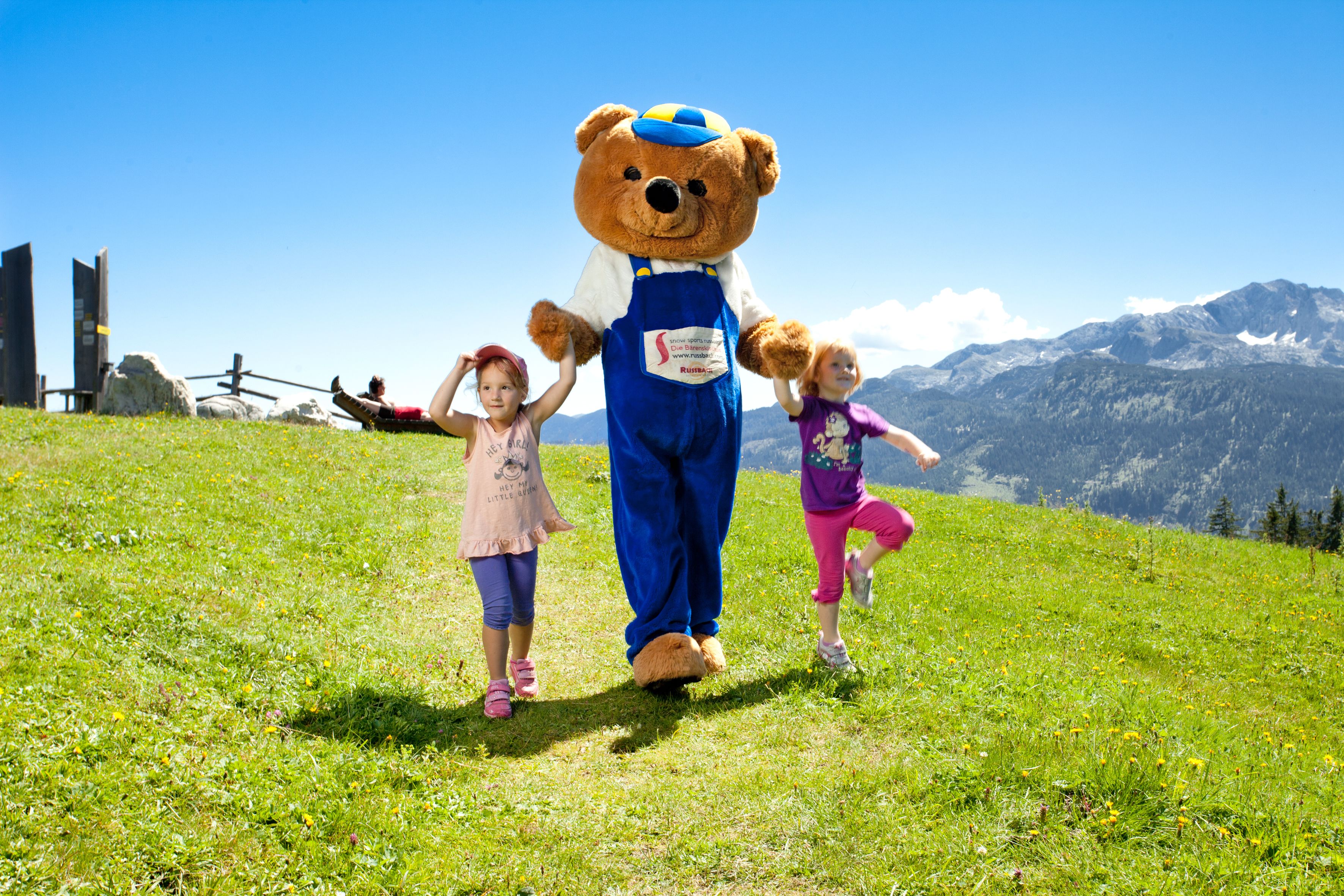 Een groot knuffelbeer-mascotte op een grasveld, met bergen op de achtergrond.