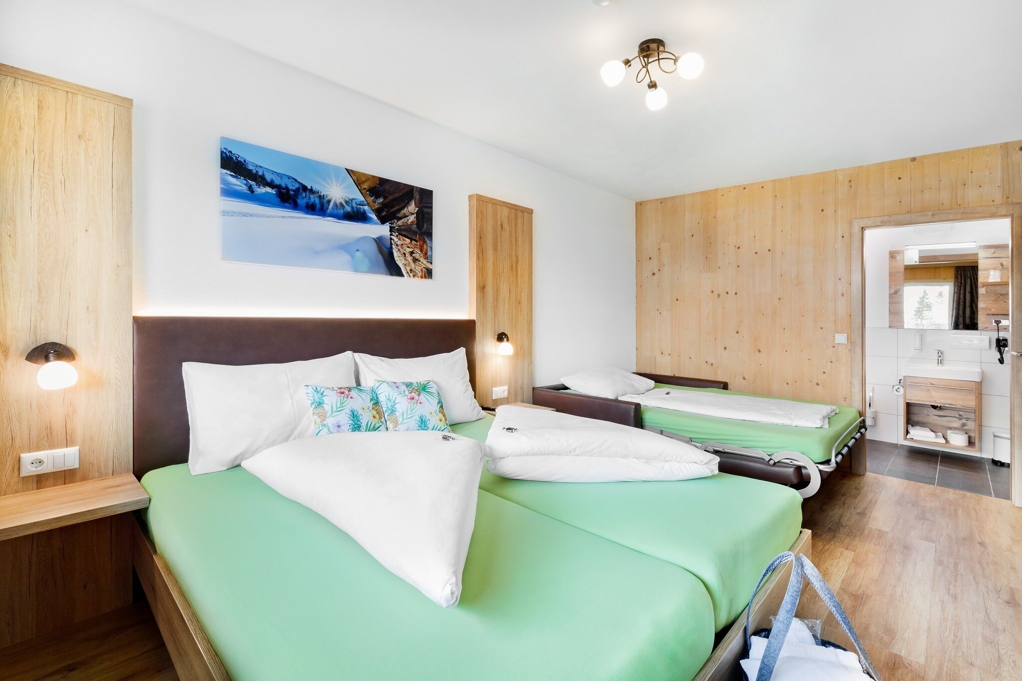 A hotel room with a large bed, a smaller bed, a wooden headboard, a painting on the wall, and a bathroom visible through an open door.