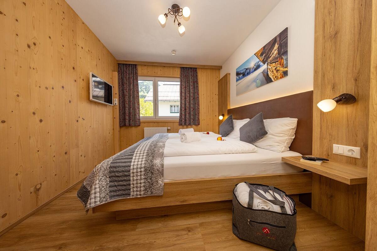 Quarto com painéis de madeira, uma cama, uma mesinha de cabeceira, uma luminária e uma janela com cortinas.