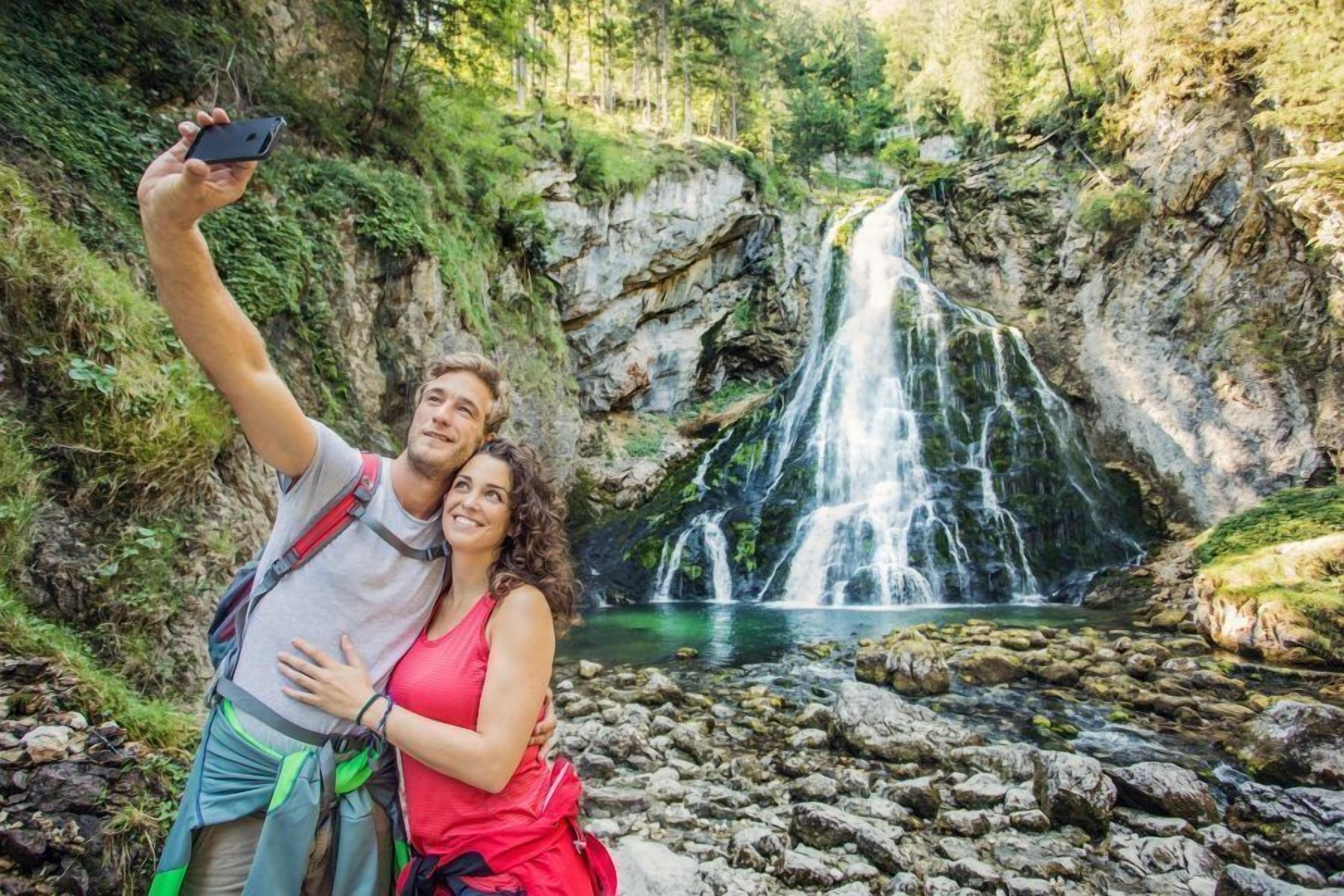 A couple taking a selfie in front of a waterfall with a rocky terrain and greenery in the background.