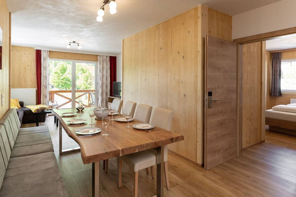 Interior de madeira com uma mesa de jantar posta para quatro pessoas, um sofá e uma cama no quarto adjacente.