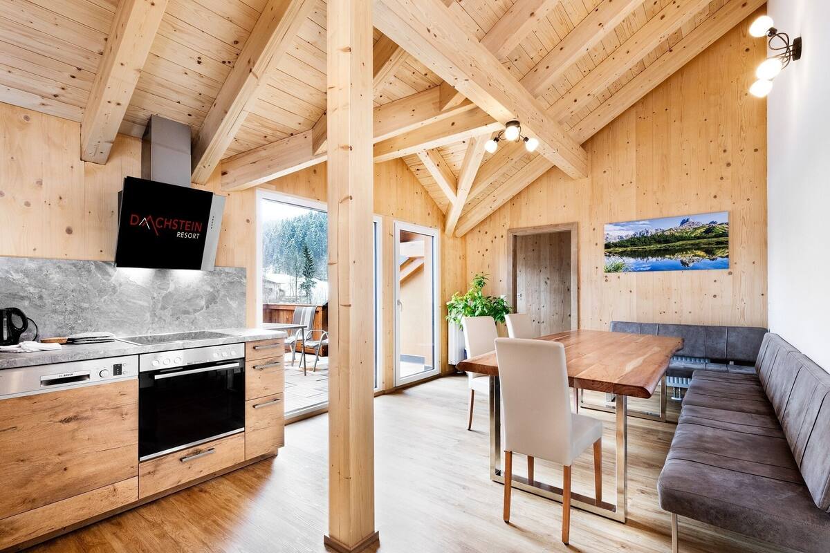 Uma cozinha moderna com forno Bosch e televisão de tela plana, vigas de madeira, área de refeições com mesa e cadeiras, e uma janela grande.