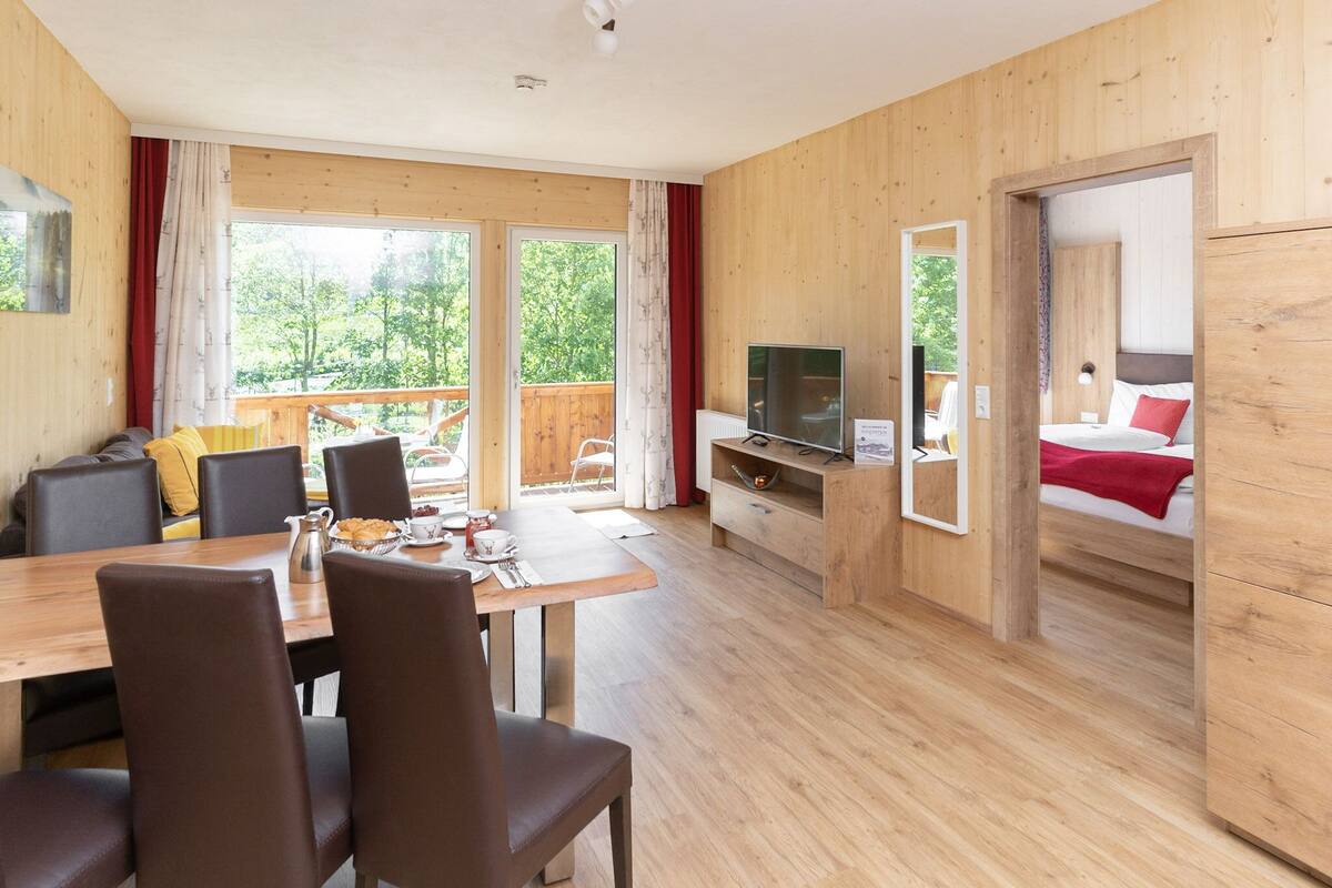 Interior em madeira com área de jantar, televisão e quarto com cama e roupa de cama vermelha.