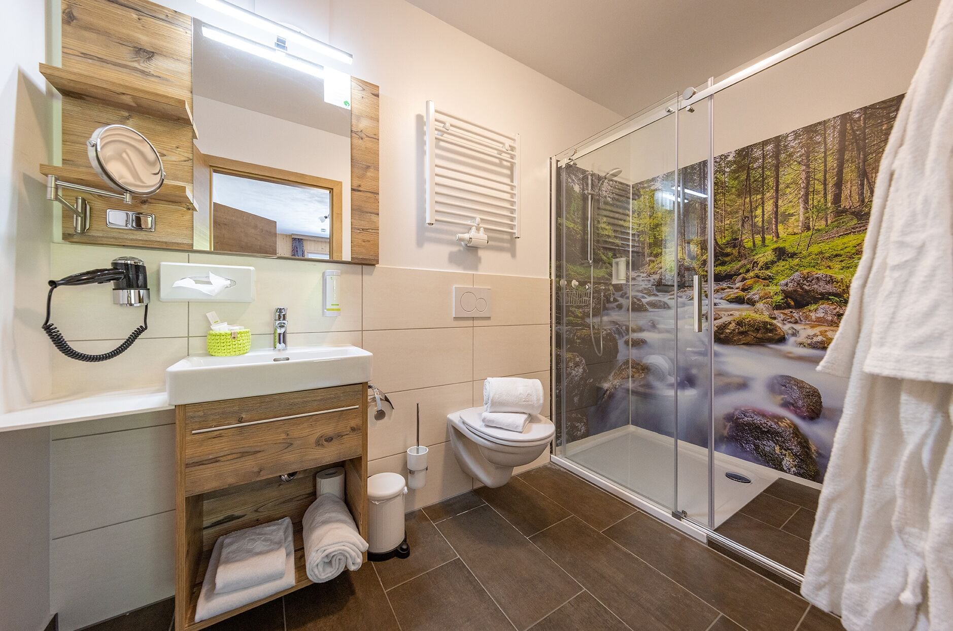 Un bagno moderno con un mobile bagno in legno, cabina doccia in vetro con vista sulla foresta e un water.