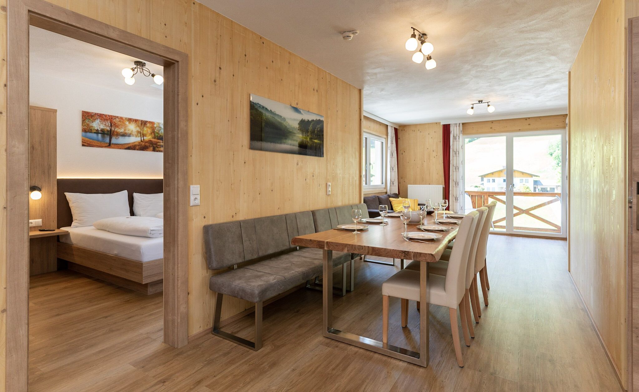 A cozy room with a bed, a bench, and a dining table with chairs. There is a painting on the wall and a window with a view.