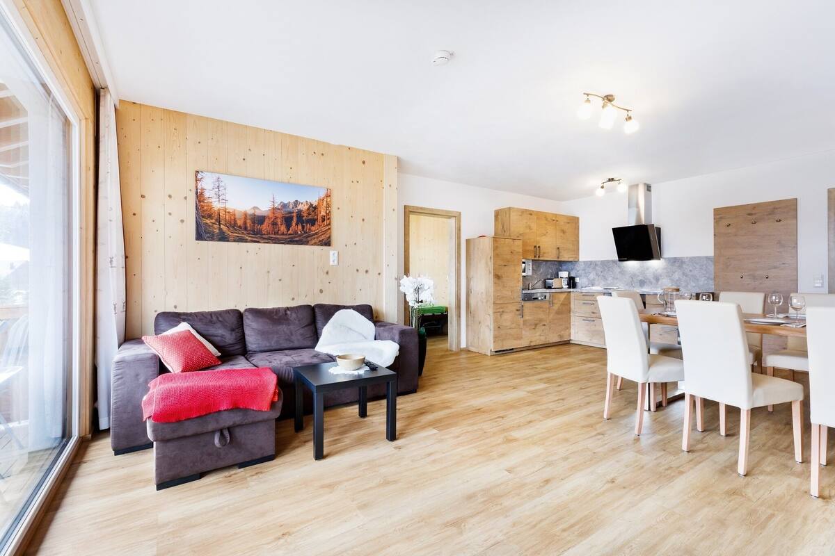 Uma sala de estar moderna com sofá, mesa de centro e área de jantar com cadeiras. Há uma cozinha com armários de madeira e uma TV de tela plana fixada acima do balcão.