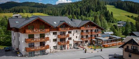 Un hotel de varios pisos con balcones, una zona de estacionamiento y vistas a un paisaje montañoso.