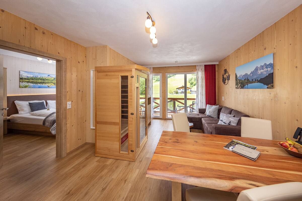 Um interior em madeira com uma cama, uma mesa de jantar com revistas e frutas, um sofá e uma janela grande com vista para as montanhas.
