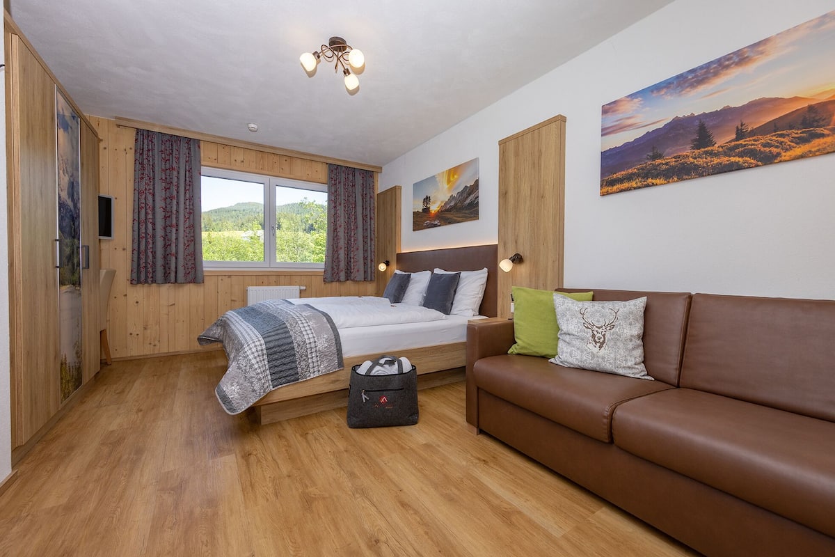 Quarto com cama, sofá de couro marrom, janela com cortinas, quadro na parede e luminária no teto.