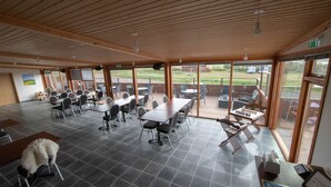 A spacious indoor area with multiple tables and chairs, large windows offering views of the outdoors, and a wooden ceiling. - Hotel Breidavik (Vesturbyggð)