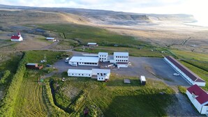 An aerial view of a rural property with multiple buildings, including a large white structure with a red roof, a smaller red-roofed building, and several smaller white buildings. There is a gravel road and a green field with a stream running through it. - Hotel Breidavik (Vesturbyggð)