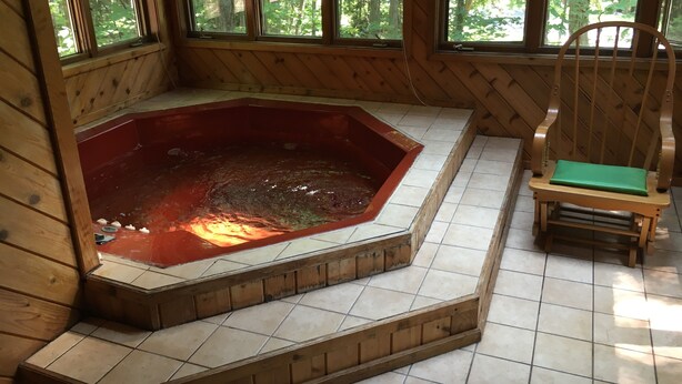 En stuga byggd av trä med ett stort, åttakantigt bubbelbad, trägolv och stora fönster med utsikt över skogen.