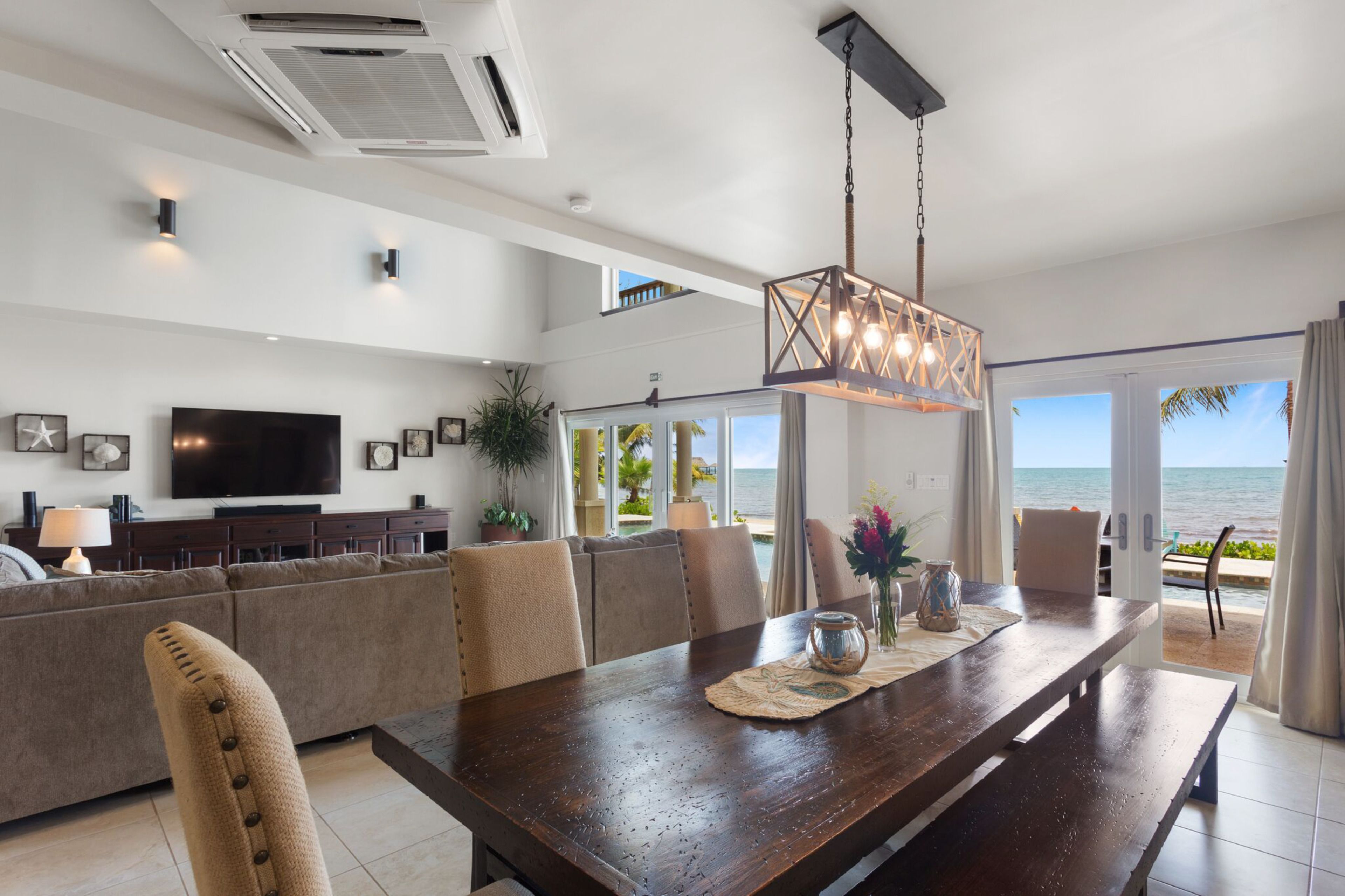 A modern living room with a large TV, comfortable seating, a wooden dining table, and a view of the ocean.