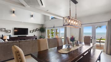 A modern living room with a large TV, comfortable seating, a wooden dining table, and a view of the ocean.