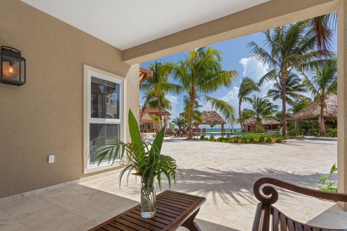 Eine Terrasse mit Holztisch und -stühlen, einem Blumentopf und Blick auf einen Strand mit Palmen und einem Gebäude mit Reetdach.