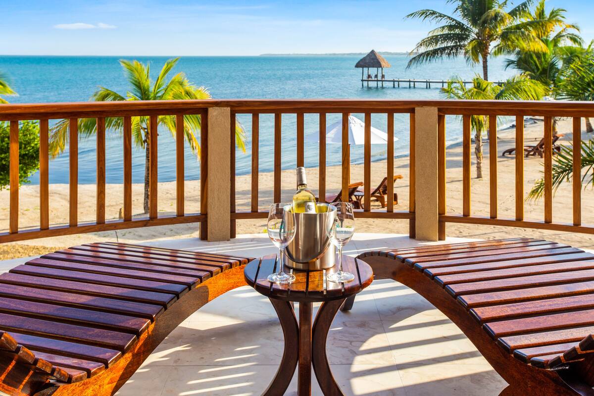 Eine Holzterrasse mit Tisch und Stühlen mit Blick auf einen Strand, an dem Palmen und ein Steg stehen.