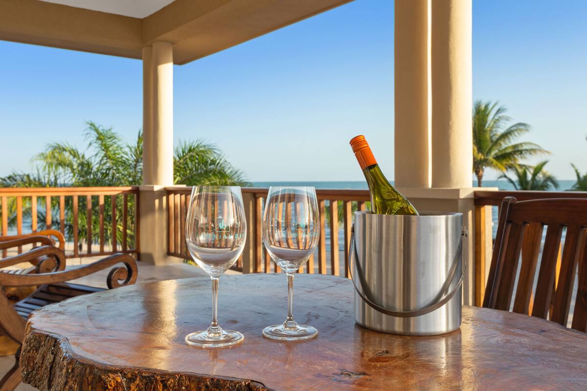 Ein Holztisch mit zwei leeren Weingläsern und einer Champagnerflasche in einem Metallkübel, platziert auf einem Balkon mit Blick auf das Meer und Palmen.