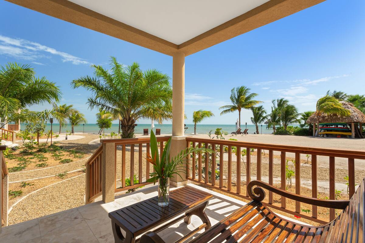 Ein Balkon mit einer Holzbank und einem Tisch, der Blick auf einen Strand mit Palmen und einem strahlend blauen Himmel bietet.