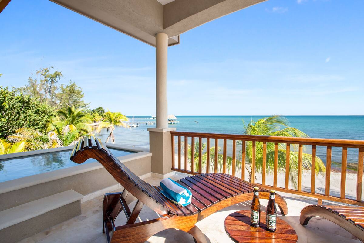 Ein Holz-Loungesessel mit Handtuch und zwei Flaschen auf einem Holztisch, mit Blick auf einen Pool und Strand mit Palmen.