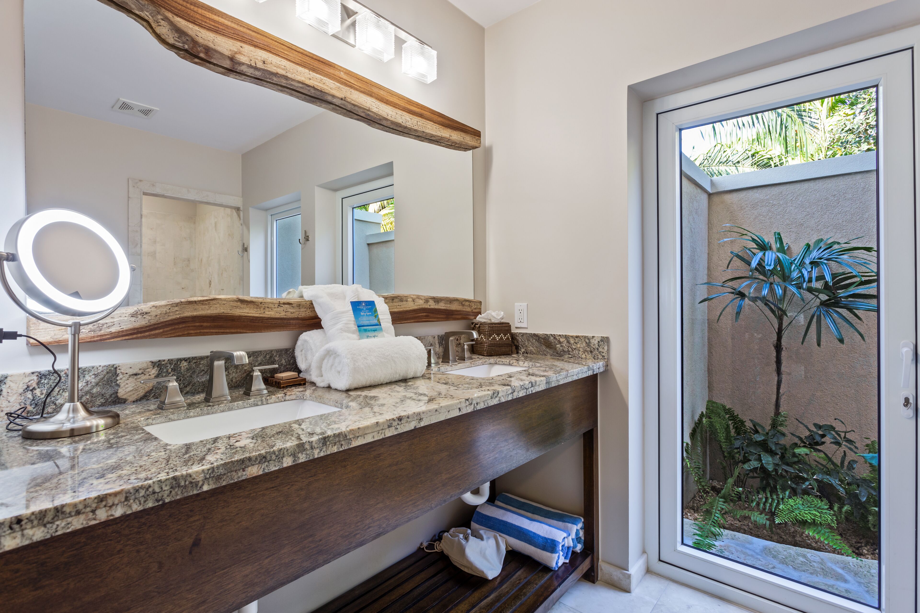 A bathroom with a marble countertop, two sinks, a large mirror with a ring light, wooden beams above the mirror, and a view of a palm tree through a glass door.