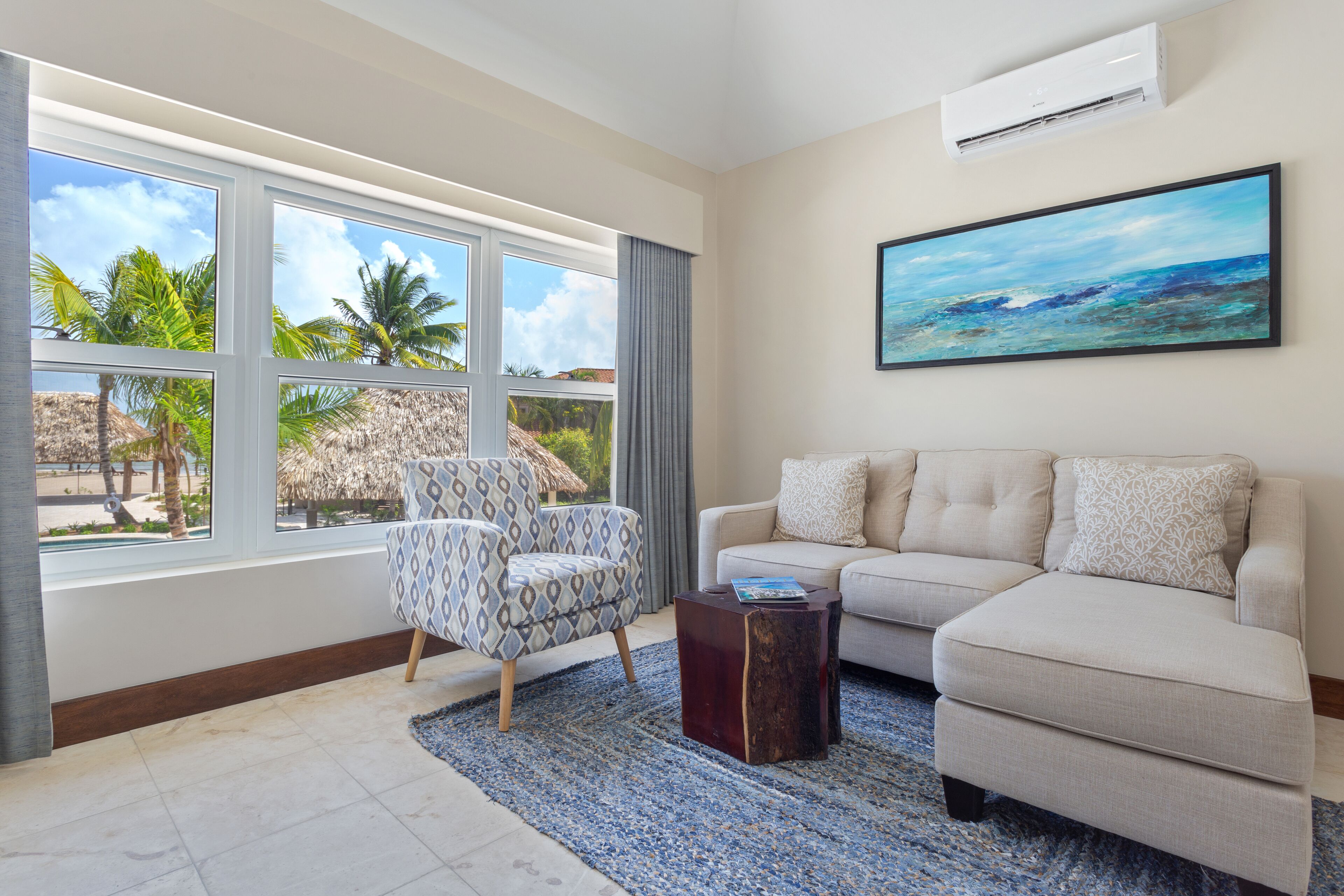Una sala de estar con sofá, sillón y un amplio ventanal con vistas a palmeras y una estructura de techo de paja.