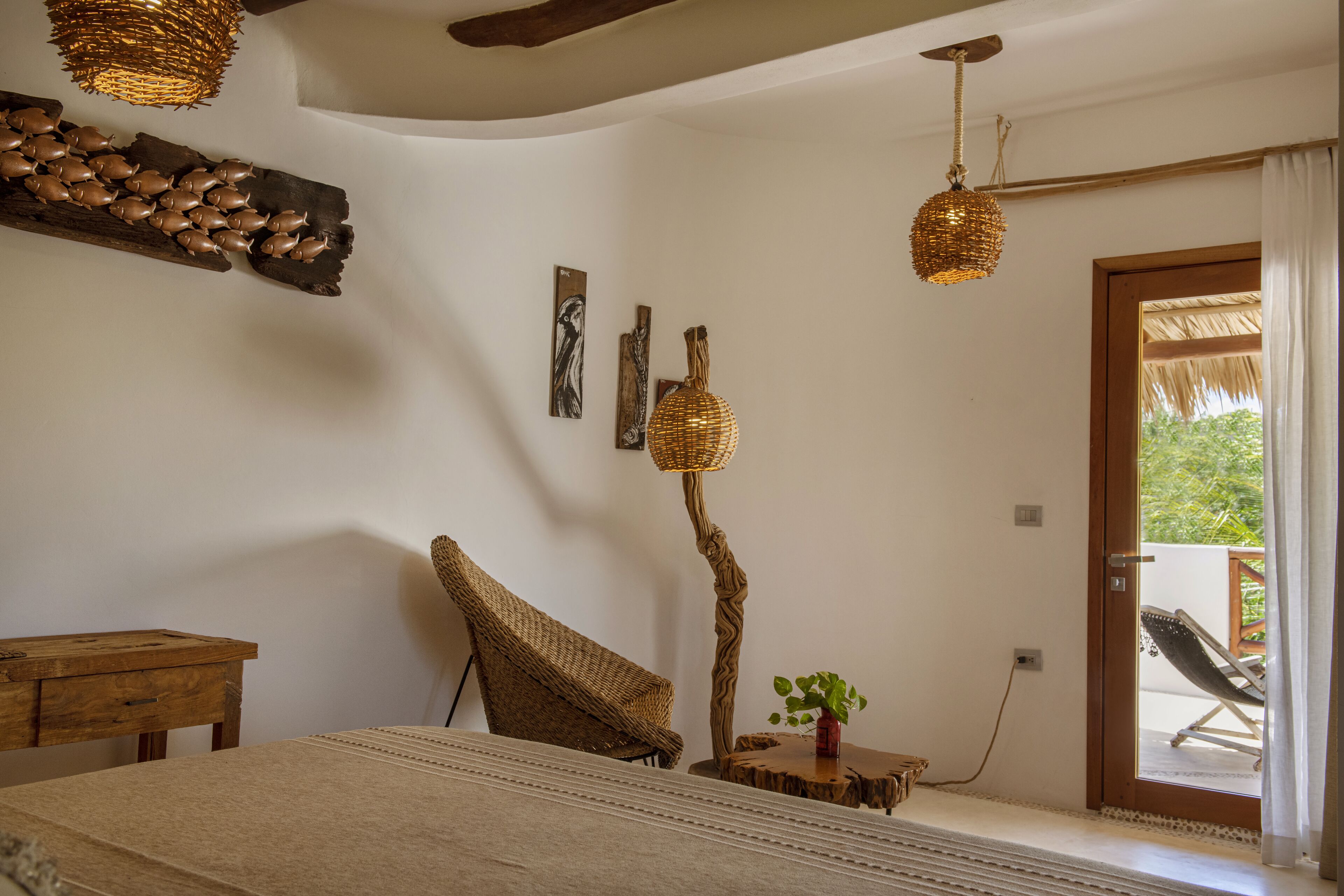 Une pièce comprenant un lit, une table en bois, une chaise en osier, un lampadaire à abat-jour en fibres tressées et une vue sur l’extérieur par une porte.