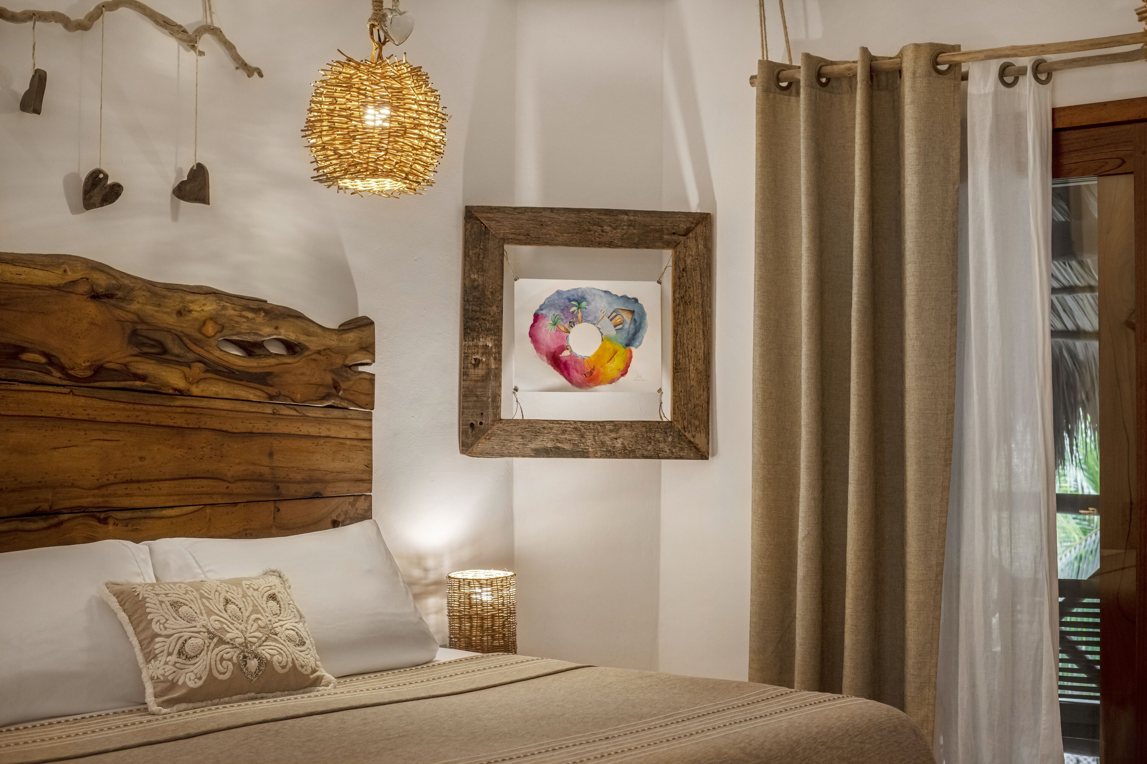 Quarto com cabeceira de madeira, cama com manta bege, obra de arte emoldurada, luminária pendente e janela com cortinas.